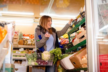 Gülümseyen kadın sebze alıyor, elinde karfiol, patlıcan, kabak ve kırmızı biber dolu sepet tutuyor..