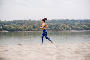 Genç bir kadın plajda koşuyor, mavi bir eşofman giyiyor ve çıplak ayakla..