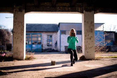 Formda bir kadın dışarıda egzersiz yapıyor, elinde tuğla tutarken çömeliyor..