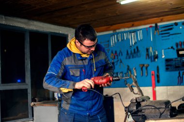 Atölyede matkap kullanan, koruyucu giysili ve gözlüklü bir adam..