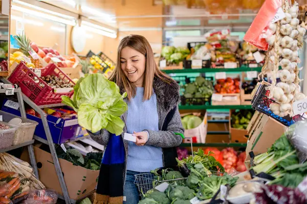 Gülümseyen güzel bir kadın manavdan yeşil salata ve dolu bir sepetle alışveriş yapıyor..