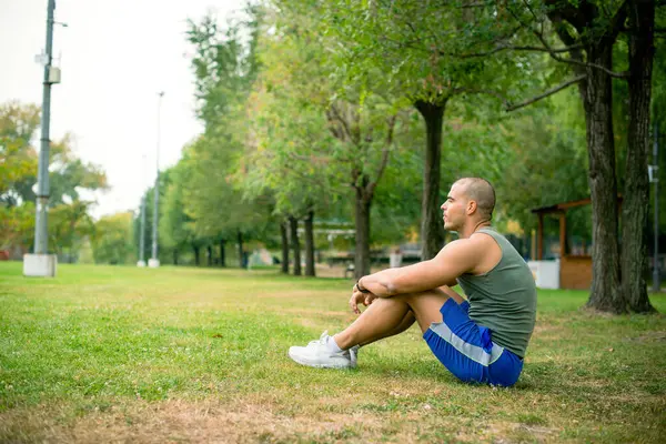 Genç sporcu sıkı spor eğitiminden sonra parkta dinleniyor, çimenlerde oturuyor..