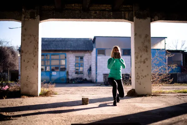 Formda bir kadın dışarıda egzersiz yapıyor, elinde tuğla tutarken çömeliyor..