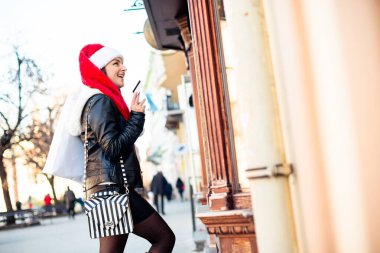 Elinde kredi kartı ve alışveriş çantasıyla dükkanın penceresinden bakarken gülümseyen genç bir kadın..