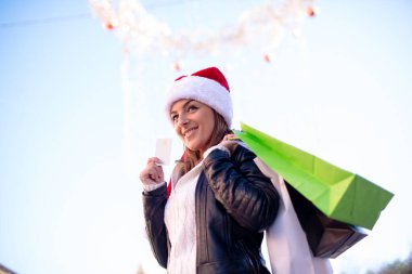 Alışveriş çantalı, elinde kredi kartı olan ve gülümseyen Noel Baba şapkalı bir kadın..