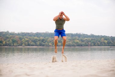 Sahilde atlayan yakışıklı erkek atlet, arka planda nehir ve açık gökyüzü..