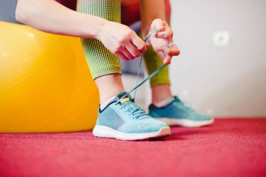 Spor salonunda ayakkabı bağlayan bir kadın, sarı bir fitness topunun üzerinde oturuyor.