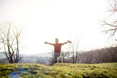 Genç adam doğada spor yapıyor, kollarını kaldırıyor ve ağırlıkları sırtına vuruyor.