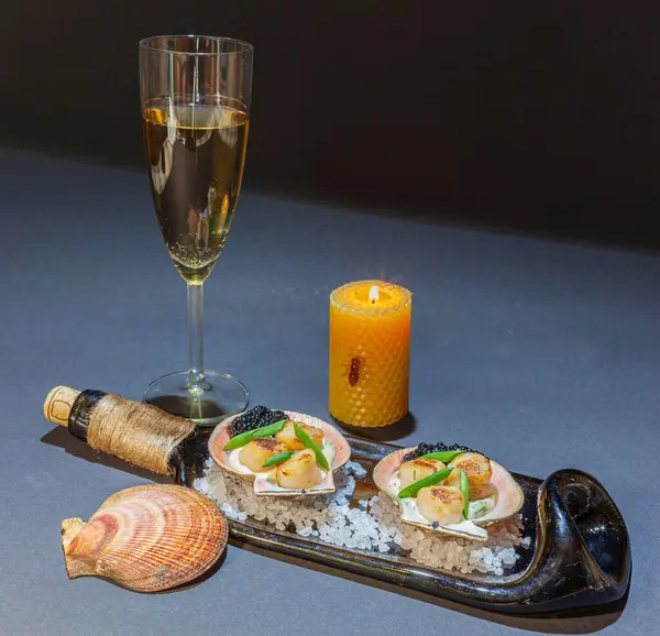 Havyarla sarımsaklı krem soslu tavada kurutulmuş istiridye, yassı şişe tabağında, balmumu mumu ve bir bardak beyaz şarap, siyah arka plan.