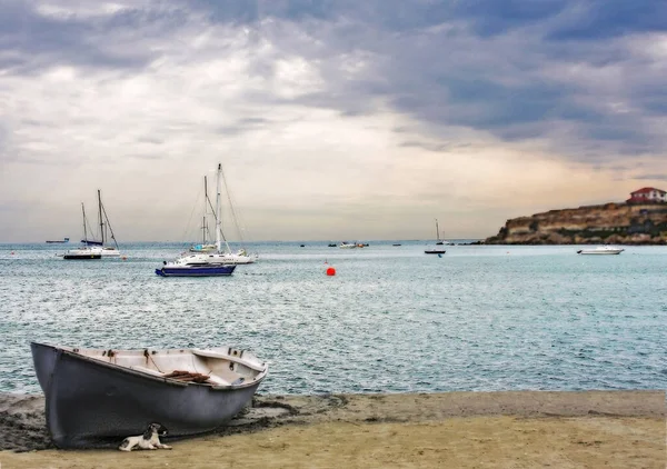 Deniz koyuna demir atmış yatlar, sakin deniz, karaya vurmuş balıkçı teknesi, bulutların arkasındaki güneş.