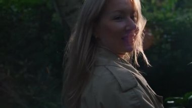 Rüzgarda saç çıkaran bir kadın. Karşıt güneş ışığında duran neşeli kadın. Rüzgar esiyor. Yeşil yaprakların etrafında