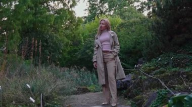 Bir kadın parkta duruyor ve başka tarafa bakıyor. Geniş açı. Açık renkli yağmurluk giyen sarışın bir kadın. Yukarıya doğru bakıyor. Etrafta ağaçlar ve yeşil yapraklar var.