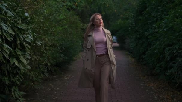 女性はカメラで動きます 周りを見回して 植物園を歩いている明るい色のレインコートのブロンドの女性 ポジティブな感情 緑の葉の周りに — ストック動画