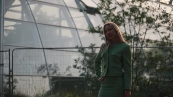 女性がカメラを歩いている 緑のスーツの金髪の女性は透明なガラスの温室の背景に対してカメラに沿って歩きます — ストック動画