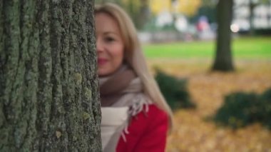 Bir kız ağacın arkasından dışarı bakıyor. Kırmızı ceketli neşeli bir kız. Sonbahar parkı. Arka planda sarı yapraklar var. Yavaş çekim