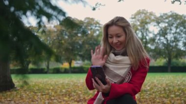 Kadın video bağlantısıyla telefonuyla iletişim kuruyor. Kırmızı ceketli bir kadın. Ağır çekim. Sonbahar parkının arka planına karşı. Aktif olarak el kol hareketi yapıyor. Elini sallıyor. Gülümsüyor