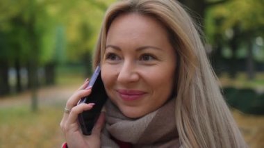 Telefonda bir kadın var. Yakın plan. Ağır çekim. Konuşurken gülümseyen ve düşünen pozitif bir kız. Elinde bir telefon tutuyordu. Arka planda sonbahar parkı.