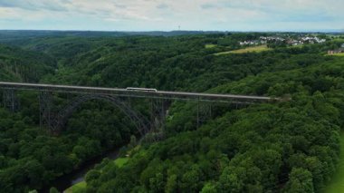 Solingen Almanya 'sında Mungstener Brucke' ye doğru yüksek irtifa drone uçuşu. Yolcu treni köprüyü geçerken dron soldan sağa doğru hareket ediyor ve Wupper Nehri, orman ve dramatik bulutlu gökyüzünü gösteriyor..
