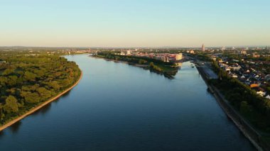 Almanya 'nın Niehl Nord Köln limanı yakınlarındaki Rhine Nehri' nde 4K insansız hava aracı uçuşu. Güneş ışığı nehri, bankalar, depolar ve binalar. Yeşillik kıyıları. Sahne, nehir kenarındaki kentsel etkinlik ve doğal güzelliğin karışımını gösteriyor..
