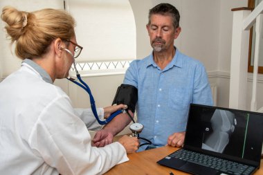 Bir doktor rutin bir randevuda hastasının tansiyonunu ölçer. Hastanın dizinin röntgeni, klinikte DR ile sohbet ederken bilgisayar ekranında gösteriliyor. Bir doktor rutin bir randevuda hastasının tansiyonunu ölçer. Hastanın röntgeni.