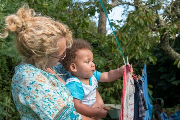 Multi-ethnic family, grandmother and grandson hang out the washing together. A biracial toddler helps with chores by handing out clothes pegs in the garden. A fun lifestyle family capture.