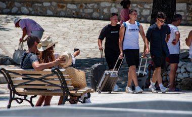 Skiathos Yunanistan. 07-05-23 Turist Bourtzi boyunca sarong 'la yürüyor. İnsanlar sıcak bir yaz gününde ağaçların gölgesinde oturdular. Yoğun bir tatil, tatil sahnesi. Boşluğu kopyala