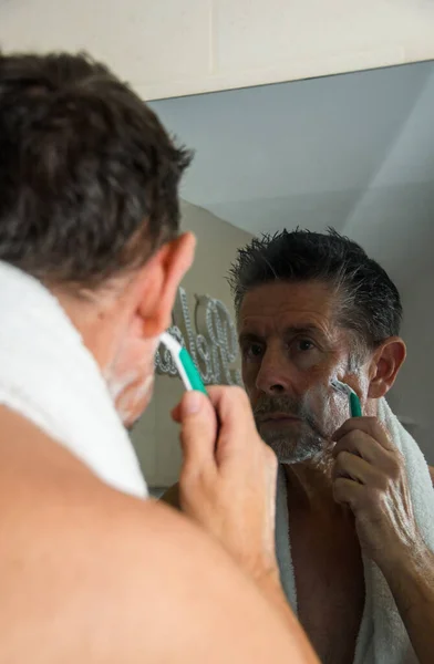 A man shaving in the mirror, his reflection is captured in a darkened bathroom.