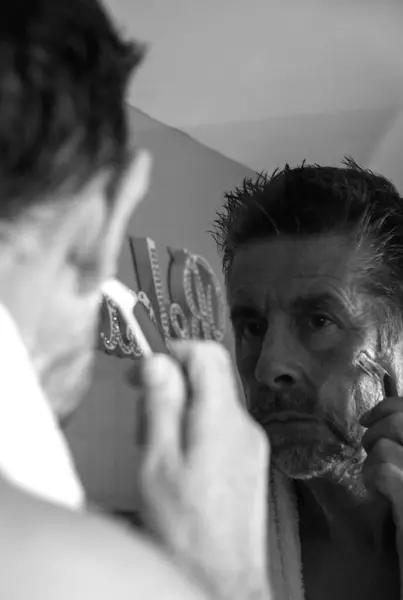 A man shaving in the mirror, his reflection is captured in a darkened bathroom. Black and white.