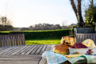 Kremalı çay, çörekler, reçel ve pıhtılaşmış krema kırsal manzaralı bir bahçe masasına konmuş. Öğleden sonra İngiliz çayı. Fotokopi alanı. Çimenli masaya odaklan, bokeh arkaplan.