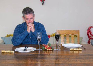 Noel imajında yalnızlık. Yanı başında boş bir sandalye ve tabakla Noel masasında tek başına oturan bir adam üzgün ve stresli görünür. Kayıp ve yalnızlık görüntüsü. Yüzüne odaklan.