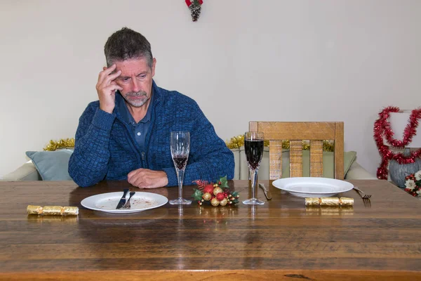 Loneliness at Christmas image. A man sits alone at a Christmas table with an empty plate by his side where someone significant would have been sat. Loss and solitude image. Selective focus on his face