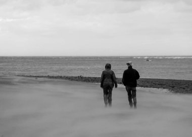 Rüzgarlı bir sahil boyunca yürüyen bir çift ayaklarının etrafında kum esiyor. Çifte odaklı siyah beyaz görüntü. Boşluğu kopyala