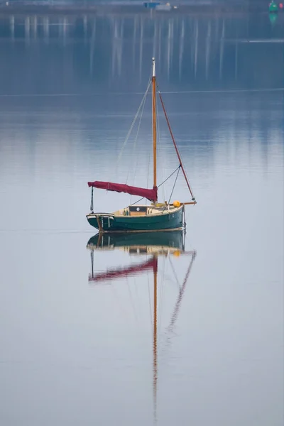A solitary sailboat sits on calm water with a clear reflection. Boat masts fill the distant background. A concept of calm solitude and isolation filled with cool blue and white colors. Boating and sea