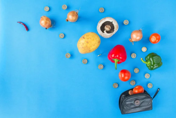 Concept image reflecting the rising cost of vegetables and food. Coins and vegetables explode out of a black purse and curl to the left. Blue background with good copy space.