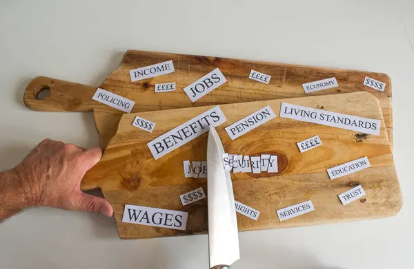Wooden chopping board with a large knife cutting up economic and social words affected by the recession and cutbacks. Wages, Benefits, living standards, Jobs etc. The knife slices through Job Security
