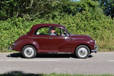 Powderham, Devon, İngiltere. 9 Temmuz 2022. Morris Minor koyu kırmızı renkli bir araba. Popüler bir meyhane arabası, bir İngiliz kır şeridinde yaz öğleden sonraları araba sürerken resmedildi. Idyllic ve ikonik tarihi araç.