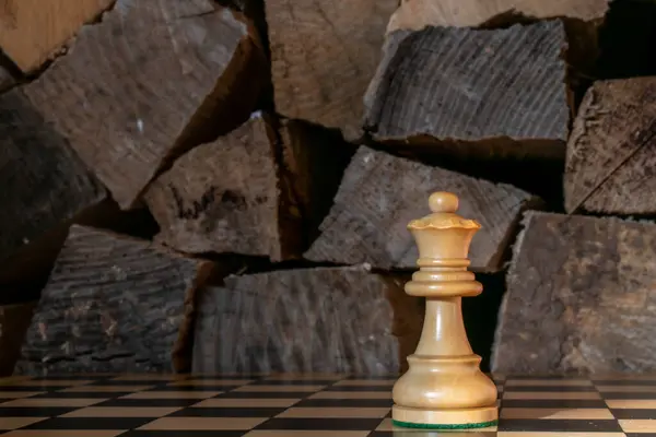 A wooden Queen chess piece stands against a backdrop of rough cut logs