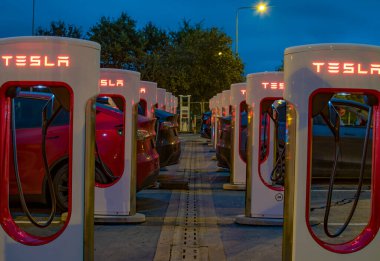 Exeter, Devon, İngiltere. 10-24-23. Tesla EV şarj istasyonları gece bir hat içinde arabalar şarj oluyor. Kırmızı Tesla işaretleri güçle parlıyor. İlk şarj istasyonuna seçici odaklan. 