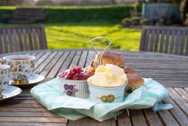 Scones with cups of tea in an English country garden. Complete with fresh Strawberry jam and Devonshire clotted cream. Dappled sunlight with green pasture beyond. Selective focus on the jam pot. clipart