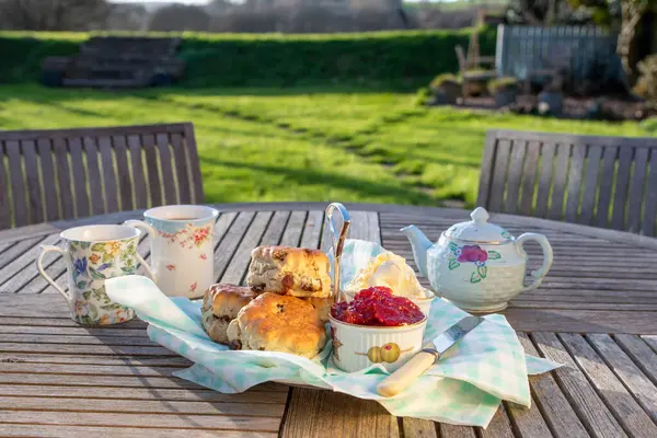 Scones with cups of tea in an English country garden. Complete with fresh Strawberry jam and Devonshire clotted cream. Dappled sunlight with green pasture beyond. Selective focus on the jam pot.