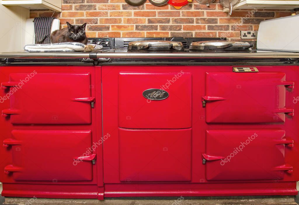 Exeter, Devon, uk. 02-10-24. Aga range cooker with a cat cuddled up on a warming plate. Country kitchen range oven in close up. Aga logo on the front. Iconic stove in an English farmhouse kitchen.