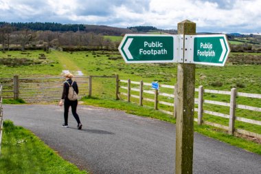 Country walks and public footpath image. A signpost marked as public footpath in sharp focus with a softly focused lady walking the path behind. Rural countryside and exercise.  clipart
