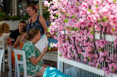 Skiathos, Yunanistan. 06-29-24. Bir kadının sıcak hava dalgası görüntüsü bir kafede oturup menüyle kendini yelpazeliyor. Kavurucu sıcaklıkta çok seksi görünüyor. Kadına odaklan..