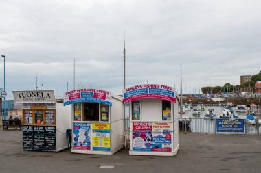 Paignton, Devon, uk. 10-06-24. Paignton harbour pleasure boats and fishing trips. Kiosks with bright signage advertising boat and fishing trips. Torbay and south devon tourism. clipart