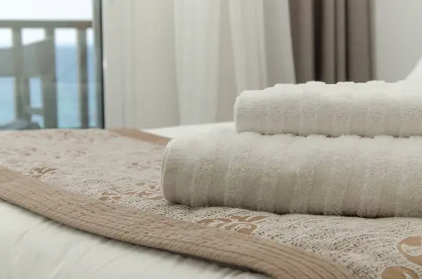 White towels lay on the bed. In the background are curtains and an open window overlooking the sea.
