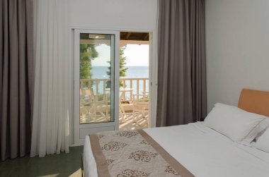 Çift kişilik yatak odası, çift kişilik yatak ve deniz manzaralı. Yatakta beyaz çarşaf, iki yastık. Yatağın kenarı kahverengi yatak örtüsüyle kaplı. Pencerenin kahverengi ve beyaz perdeleri var. Yunanistan, Halkidiki, Sithonia, Elia.