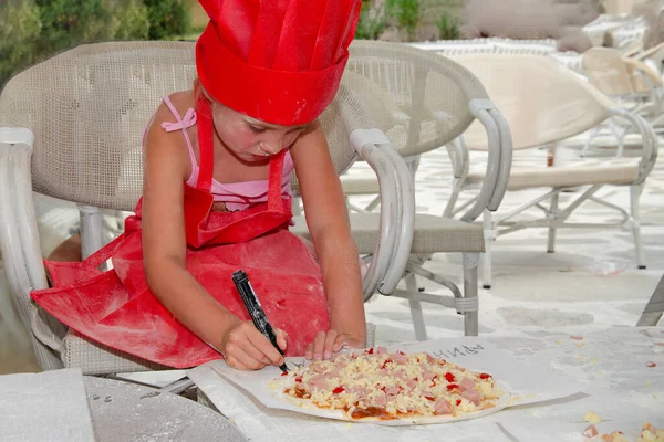 Five year old girl in red chef cap apron apron makes pizza. She is on summer terrace with white chairs and table