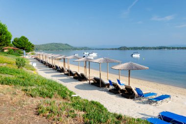 Bol bol güneş şezlongları ve şemsiyeleri olan temiz bir sahil. Denizde kayıklar var. Ön tarafta çim var.