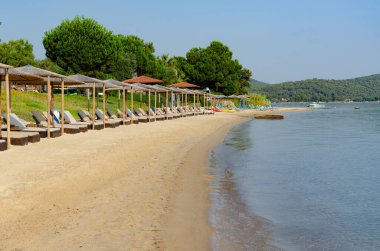 Bol bol güneş şezlongları ve şemsiyeleri olan temiz bir sahil. Denizde kayıklar var. Ön tarafta çim var.