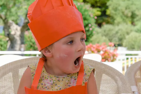 five-year-old girl yawns in cooking class. She is wearing a chefs hat and apron
